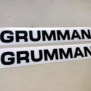 May include: Two rectangular white stickers with the word "GRUMMAN" printed in bold, black letters. The stickers are placed side by side against a light gray background. The text is centered on each sticker.