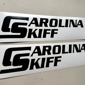 May include: Two white rectangular stickers with the words "CAROLINA SKIFF" in bold black letters. The "S" in "SKIFF" is stylized and overlaps the word "CAROLINA". The stickers are on a light gray surface.