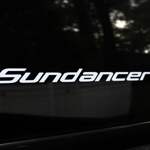 May include: White "Sundance" decal on a black vehicle window. The decal is in a stylized font, with the letters slightly angled. The background is blurred, showing trees and a glimpse of the sky.