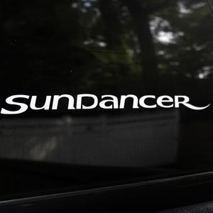 May include: White vinyl decal with the word "SunDancer" in a stylized font, applied to a black surface. The decal is likely for a vehicle or window, with a curved flourish at the end of the word.