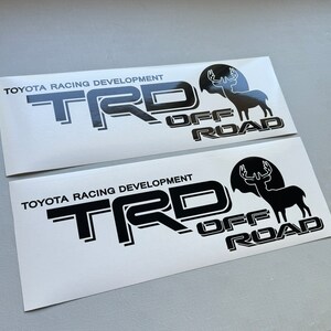 Puede incluir: Dos pegatinas rectangulares blancas con texto y gráficos negros. El texto dice "TOYOTA RACING DEVELOPMENT" encima de las letras "TRD" y "OFF ROAD". Una silueta negra de un ciervo con cuernos está en el lado derecho de la pegatina.