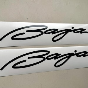 May include: Two white rectangular stickers with the word "Baja" in a black, cursive font. The stickers are placed side by side, with the text running horizontally across the center of each sticker. The background is a light grey.