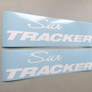 以下が含まれることがあります： 白字で「Sur TRACKER」と書かれた、2つの水色の長方形ステッカー。「Sur」は筆記体、「TRACKER」は太字のサンセリフ体です。ステッカーは車両用です。