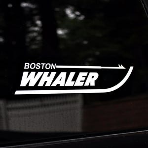 Puede incluir: Calcomanía de vinilo blanco con las palabras "BOSTON WHALER" en negrita, fuente sans-serif. La calcomanía presenta una flecha estilizada y un diseño de línea curva. La calcomanía está en una superficie negra.