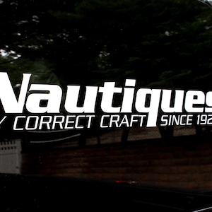 May include: White decal on a black surface. The decal reads "Nautiques" in a bold, stylized font, with "BY CORRECT CRAFT" below and "SINCE 1925" at the bottom. The background is blurred, suggesting an outdoor setting.