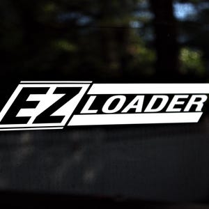 Peut inclure: Autocollant blanc "EZ LOADER" sur fond noir. Le texte est en gras, sans empattement, avec "EZ" dans un design stylisé et angulaire. Le mot "LOADER" est en ligne droite horizontale.