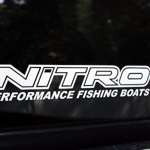 May include: White decal with the word "NITRO" in a bold, outlined font, above the words "PERFORMANCE FISHING BOATS". The decal is affixed to a dark surface, likely a vehicle window.