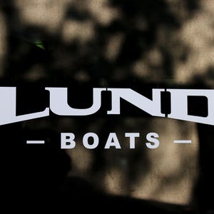 May include: White text on a dark background reads "LUND BOATS". The text is centered, with "LUND" in a bold, stylized font above "BOATS". Dashes are on either side of "BOATS".