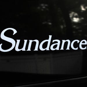 May include: White "Sundance" decal on a black surface. The word is in a cursive font, with the letters slightly connected. The background is dark, with hints of trees visible.