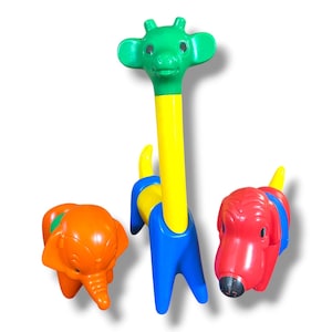 Può includere: Tre giocattoli animali in plastica che includono un elefante arancione con una fascia verde, una giraffa alta con testa verde, collo giallo e corpo blu, e un cane rosso con una fascia blu e coda gialla, tutti con superfici modellate lisce.