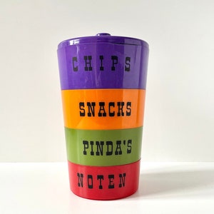 May include: A multi-colored plastic container with four sections. The sections are labeled "CHIPS", "SNACKS", "PINDAS", and "NOTEN".