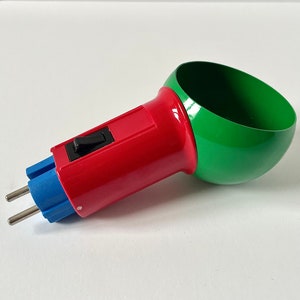 Peut inclure: Un adaptateur de prise électrique rouge et vert avec un interrupteur noir. L'adaptateur a deux broches et un connecteur en plastique bleu.