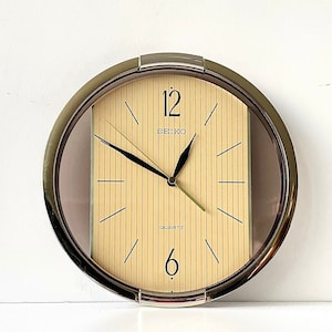 May include: A round, gold-toned wall clock with a beige face and black hands. The clock has a glass cover and the words "SEIKO QUARTZ" printed on the face.