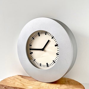 May include: A silver round clock with black hands and black numbers on a white face. The clock is sitting on a wooden surface.