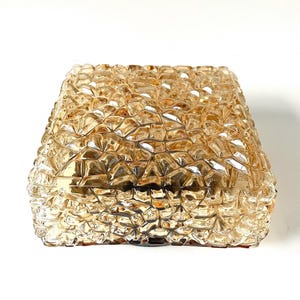 May include: A square, amber-colored glass light fixture with a textured, geometric pattern. The fixture has a faceted surface, creating a sparkling effect. The design is reminiscent of cut crystal or a mosaic.