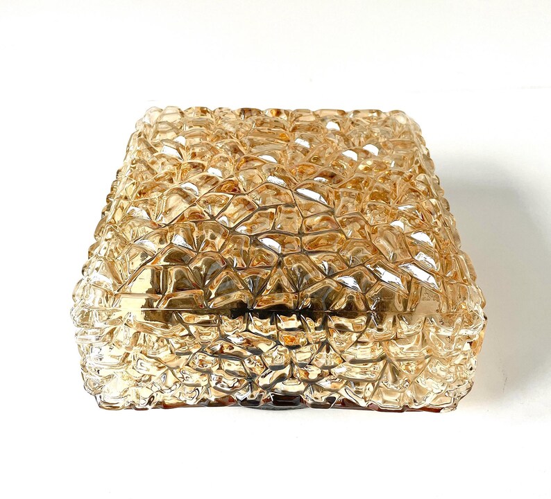 May include: A square, amber-colored glass light fixture with a textured, geometric pattern. The fixture has a faceted surface, creating a sparkling effect. The design is reminiscent of cut crystal or a mosaic.