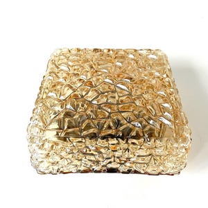 May include: A square, amber-colored glass light fixture with a textured, geometric pattern. The fixture has a slightly tapered shape and is designed to be mounted on a wall or ceiling. The glass has a warm, golden hue.