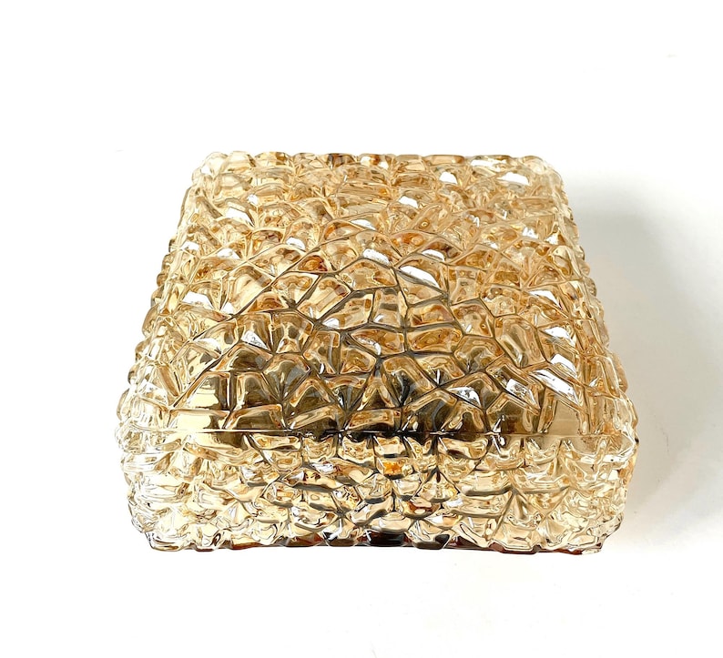 May include: A square, amber-colored glass light fixture with a textured, geometric pattern. The fixture has a slightly tapered shape and is designed to be mounted on a wall or ceiling. The glass has a warm, golden hue.