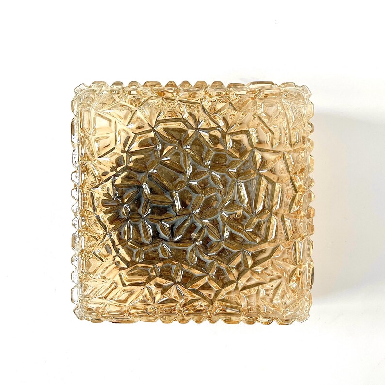May include: A square, amber-colored glass light fixture with a textured, geometric pattern. The fixture has a slightly rounded edge and a warm, inviting glow. The design is reminiscent of vintage or retro styles.