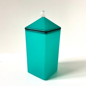 Könnte beinhalten: Eine tealblaue Glasdose mit Deckel und einem durchsichtigen Glasknopf oben. Die Dose hat einen schwarzen Rand oben.