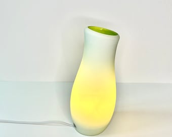 May include: A modern, illuminated table lamp with a curved, vase-like shape. The lamp features a gradient color scheme, transitioning from white to yellow and green at the top. The lamp is plugged in and emits a warm, inviting glow.