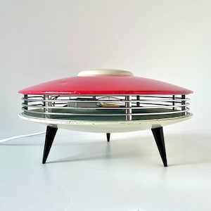 May include: A red and white mid-century modern table lamp with a circular base and three black legs. The lamp has a chrome wire cage around the light bulb.
