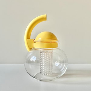 May include: A clear glass teapot with a rounded body and a yellow plastic lid. The teapot features a tall, curved yellow handle attached to the side and a central, cylindrical, perforated clear plastic infuser basket hanging from the lid.