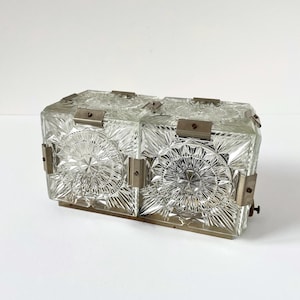 May include: A rectangular wall light fixture featuring two clear glass blocks with a pressed starburst pattern. The glass sections are held together by brushed metal clips and a base plate, creating a structured, geometric lighting design.