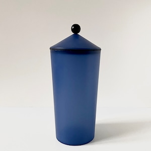 Könnte beinhalten: Eine blaue Keramikdose mit einem schwarzen Knopf auf dem Deckel. Die Dose ist hoch und zylindrisch.