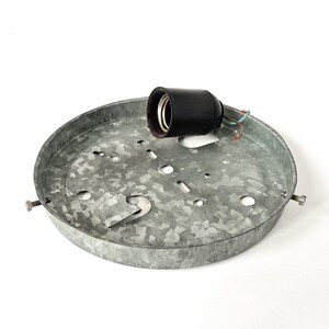 May include: A galvanized metal ceiling light fixture with a black light socket. The fixture is round with several holes and a bracket for mounting. The light socket has exposed wires. The fixture is designed for indoor lighting.