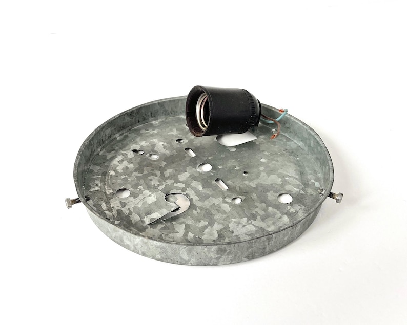 May include: A galvanized metal ceiling light fixture with a black light socket. The fixture is round with several holes and a bracket for mounting. The light socket has exposed wires. The fixture is designed for indoor lighting.