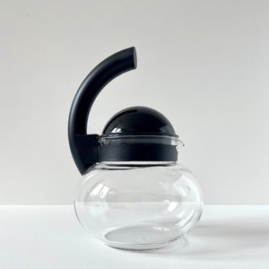 Può includere: Una caraffa per caffè in vetro trasparente con corpo rotondo e manico e coperchio neri. Il manico è curvo verso l'alto e il coperchio ha una forma arrotondata. La caraffa è progettata per preparare il caffè.