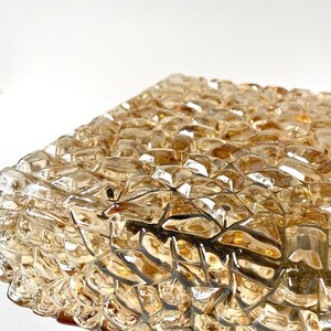 May include: Close-up of a textured, amber-colored glass object. The surface is composed of numerous irregular, geometric shapes, creating a mosaic-like appearance. The glass reflects light, highlighting its multifaceted design.