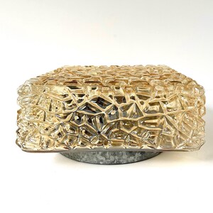 May include: A square, amber-colored glass light fixture with a textured, geometric pattern. The fixture sits on a circular, gray metal base. The glass has a faceted appearance, creating a unique lighting effect.