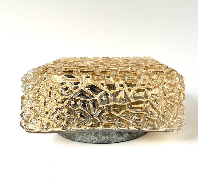 May include: A square, amber-colored glass light fixture with a textured, geometric pattern. The fixture sits on a circular, gray metal base. The glass has a faceted appearance, creating a unique lighting effect.