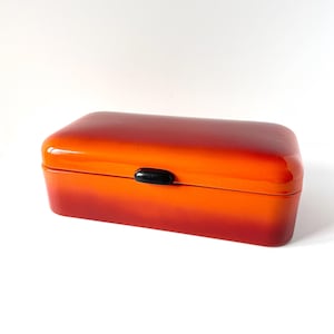 May include: A rectangular storage box with a smooth, glossy finish that transitions from a bright orange top to a deep red base. The container features a rounded lid and a small, centered black oval latch on the front for secure closure.