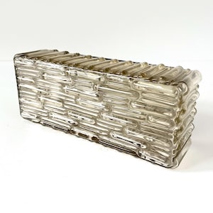 May include: A rectangular, clear glass decorative box with a textured, brick-like pattern. The box has a slightly tinted, smoky appearance and is designed with a series of horizontal and vertical lines, creating a woven effect. The glass is thick and sturdy.
