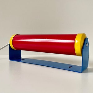 May include: A red and yellow cylindrical lamp with a blue metal base. The lamp has a white cord and is mounted on a white wall.