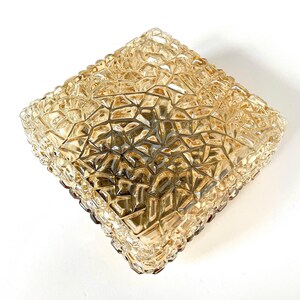 May include: A square, amber-colored glass ceiling light fixture with a textured, geometric pattern. The fixture has a faceted surface and a brown border. The light is designed to be mounted on a ceiling and provides ambient lighting.