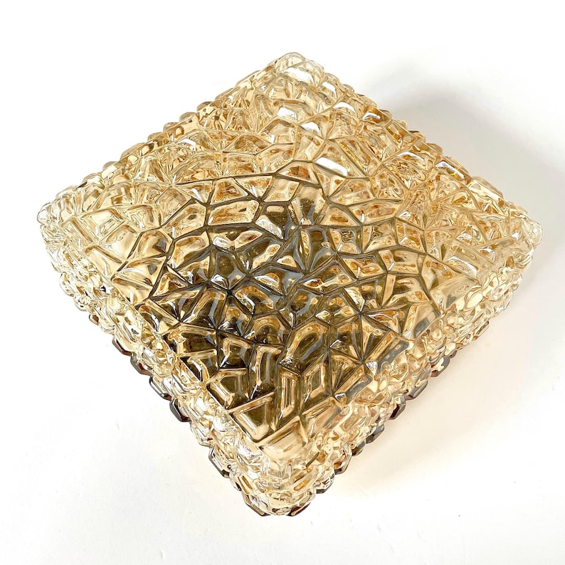 May include: A square, amber-colored glass ceiling light fixture with a textured, geometric pattern. The fixture has a faceted surface and a brown border. The light is designed to be mounted on a ceiling and provides ambient lighting.