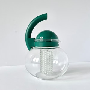 Może przedstawiać: Przezroczysty szklany czajnik o kulistym korpusie i ciemnozielonym uchwycie i pokrywce. Czajnik posiada cylindryczny zaparzacz w środku. Uchwyt zakrzywia się do góry, a następnie w dół. Pokrywka ma zaokrąglony kształt kopuły.