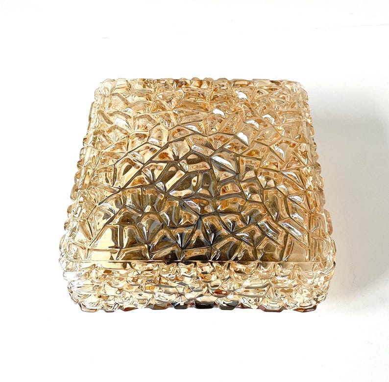May include: A square-shaped, amber-colored glass lampshade with a textured, geometric pattern. The shade has a slightly flared shape and a warm, inviting glow. The design is reminiscent of cut crystal or faceted glass.