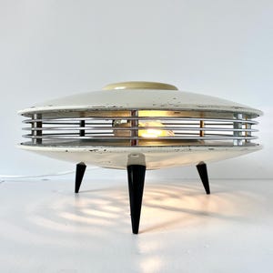 May include: A vintage, cream-colored table lamp with a retro, space-age design. The lamp features a circular shade with horizontal chrome bars and three black, conical legs. The lamp is lit, casting a warm glow.