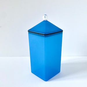 Può includere: Un contenitore alto, azzurro con una forma geometrica e un pomello sferico trasparente sul coperchio. Il contenitore ha una base quadrata e un corpo leggermente affusolato. Il coperchio è di una tonalità di blu più scura e ha un bordo nero.