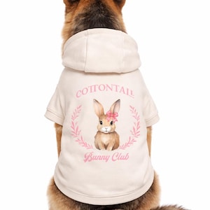 May include: A dog wearing a cream-colored hoodie with a bunny graphic. The hoodie features the text "Cottontail Bunny Club" in pink, along with a watercolor illustration of a bunny wearing a pink bow. The dog is a German Shepherd.