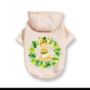 May include: A light beige dog hoodie with a hood. The back features a St. Patrick's Day design with a wreath of green shamrocks, a yellow bow, and a pot of gold. The word "PETSIN" is printed on the hood.