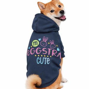 May include: A Shiba Inu dog wearing a navy blue hoodie with the text "I'm Eggstra Cute" in colorful lettering. The hoodie features bunny graphics and heart accents, perfect for Easter or a fun pet outfit.
