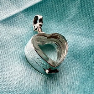 Op de afbeelding: Een zilveren hartvormig medaillon met een transparante glazen inzet. Het medaillon heeft een inscriptie met de tekst "Joshua".