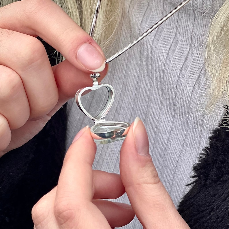 May include: A silver-toned heart-shaped locket pendant attached to a thin snake chain. The locket is open, revealing a hollow interior space. The metal has a polished, reflective finish, and the design features a simple, clean structure.