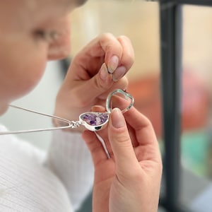 Op de afbeelding: Een zilveren ketting met een hartvormige hanger en een transparant glazen deksel. De hanger is gevuld met kleine paarse amethistkristallen.